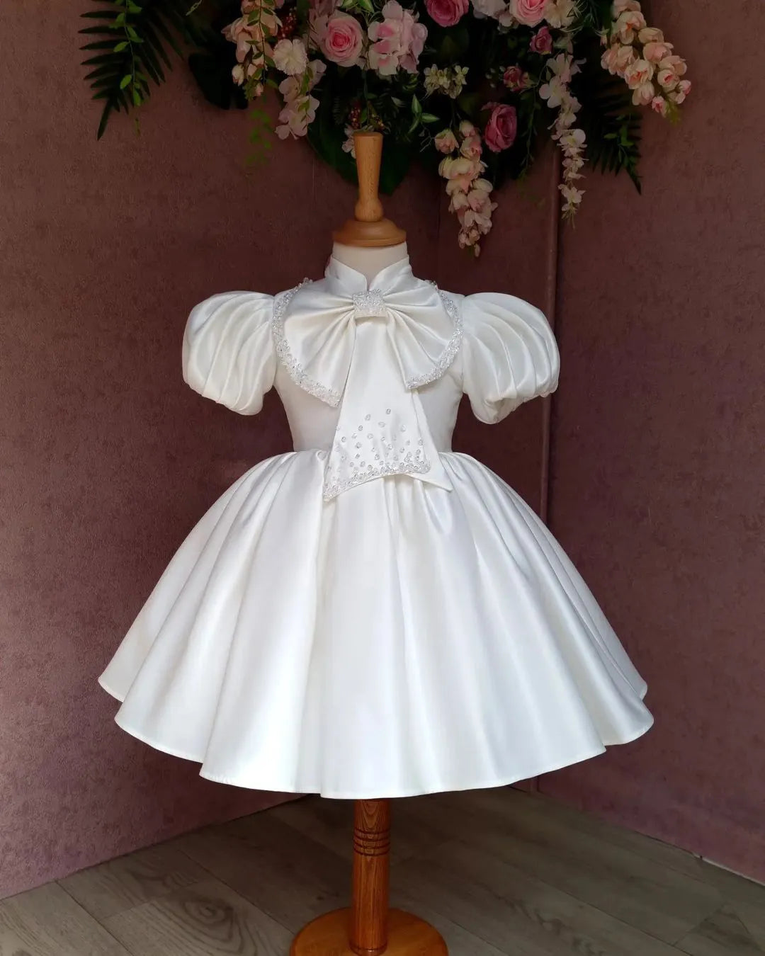  Front view of an ivory satin dress for girls with a large, embellished bow at the front.